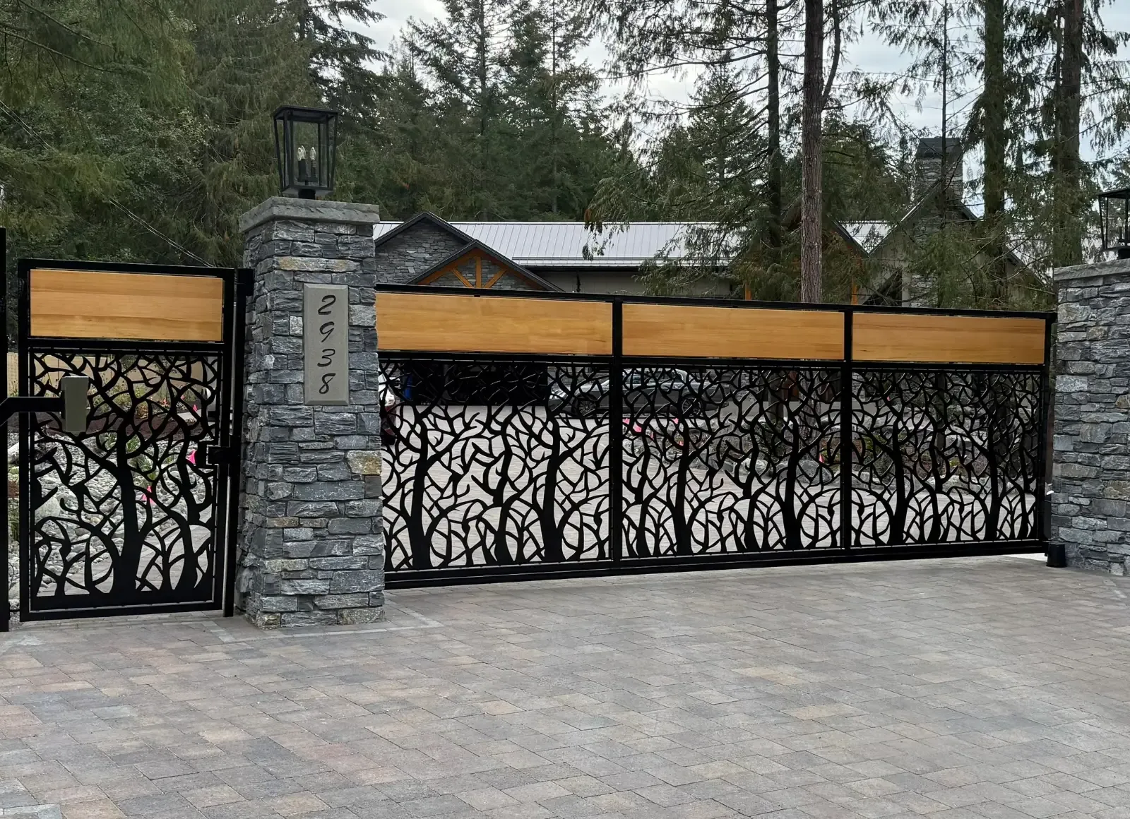 Sliding driveway gate and fence with laser-cut forest panels, cedar top rail and stone pillars